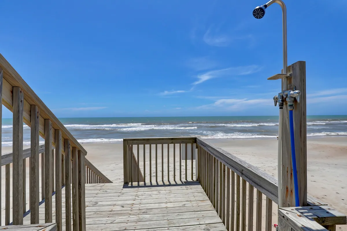 A wooden walkway leads from the property toward the shore, featuring an outdoor shower and an open ocean view. The path is inviting, emphasizing the ease of beach access and the natural beauty surrounding the property. The openness of the scene highlights space for both people and pets to enjoy outdoor play and post-beach clean-ups.