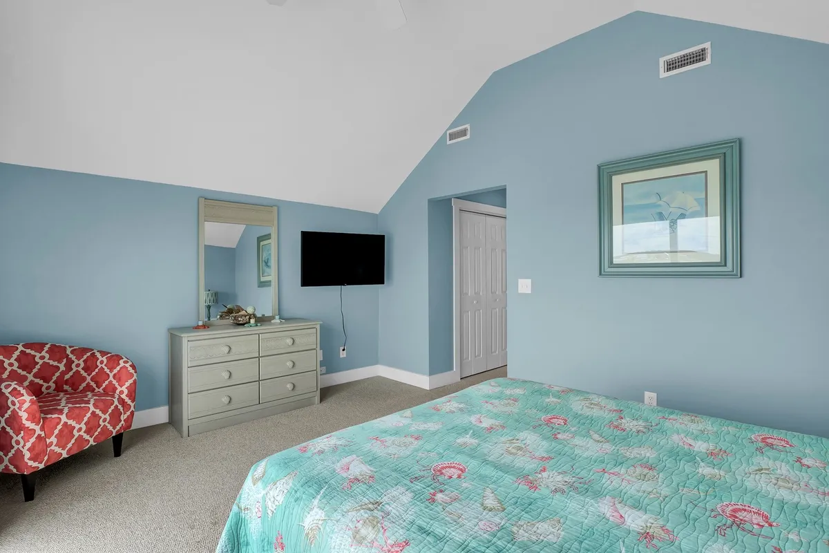 A cozy bedroom featuring a bed with a colorful quilt, a red patterned chair, a mirror, and a mounted TV, offering a relaxing and comfortable environment ideal for rest and privacy.