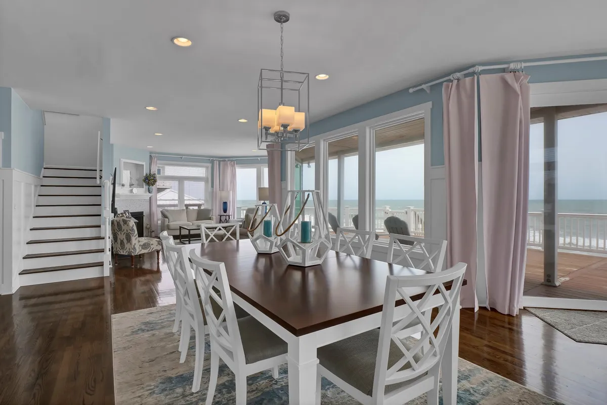 A bright dining area with a large table, stylish chandelier, comfortable seating, and expansive windows offering ocean views. The layout is open and airy, ideal for both special occasions and everyday meals, with the ocean visible from every seat.