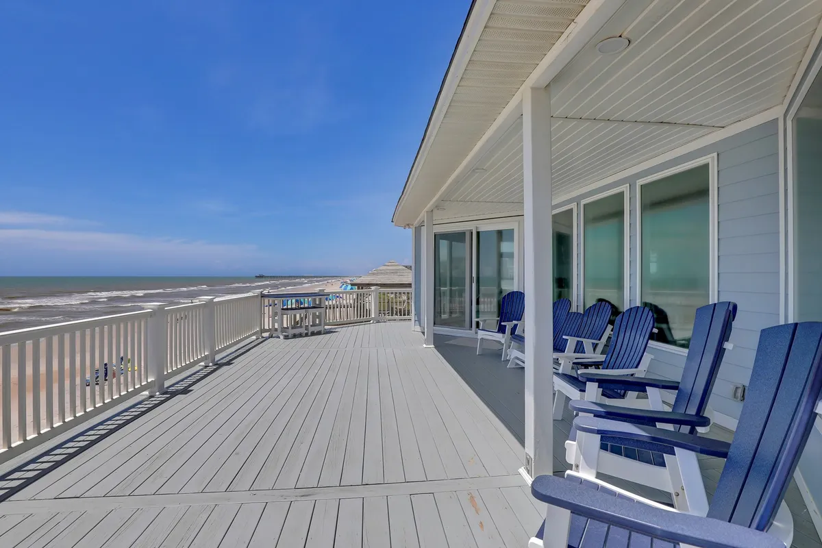 An expansive deck positioned directly above the beach with comfortable chairs facing a breathtaking ocean view and blue sky. The deck's elevated vantage point captures the open water and horizon, with sunlight casting a serene glow on the scene, suggesting a tranquil retreat for relaxation or socializing outdoors.
