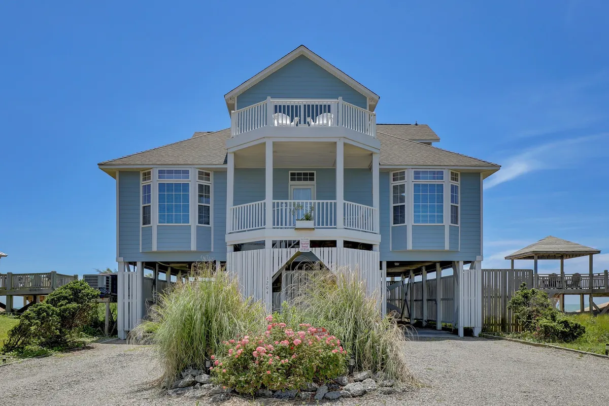 This exterior image showcases the main entrance of the home, a spacious wrap-around balcony, and a landscaped garden. The setting is cheerful and inviting, hinting at possibilities for group gatherings and outdoor activities.