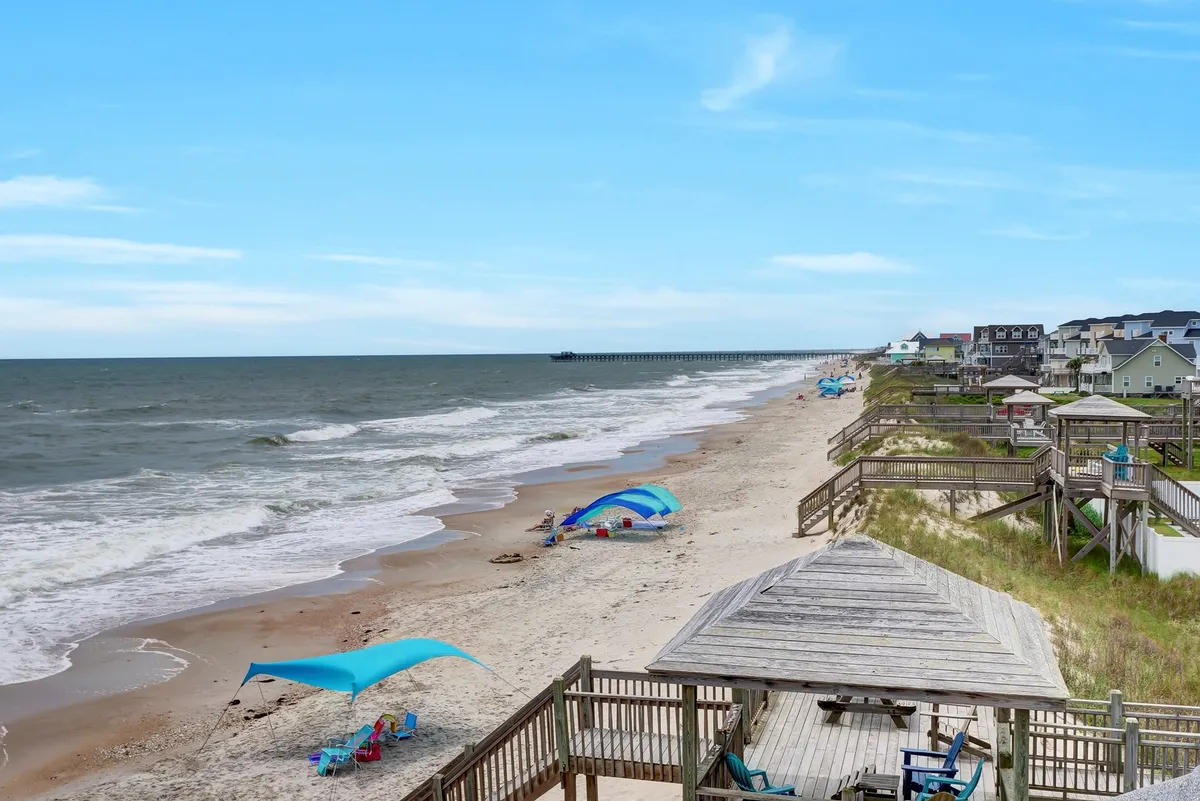 A classic beach scene with colorful umbrellas, gentle ocean waves, a long boardwalk, and a distant pier. Children and families can be imagined building sandcastles, swimming, and enjoying the sun.