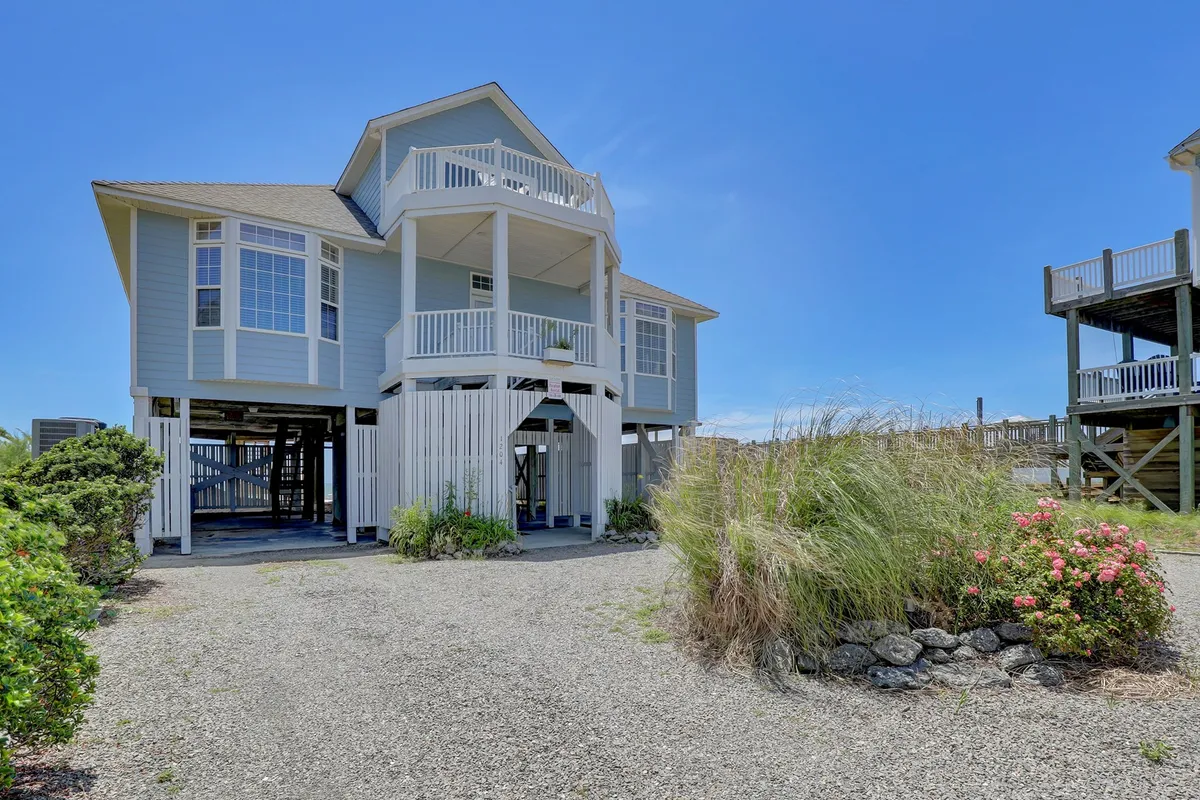 A welcoming exterior shot featuring large windows and a wrap-around porch bordered by a garden area. The porch is ideal for gatherings, stargazing, or simply soaking in the surroundings, while the architectural features express elegance and a close relationship with the outdoors.