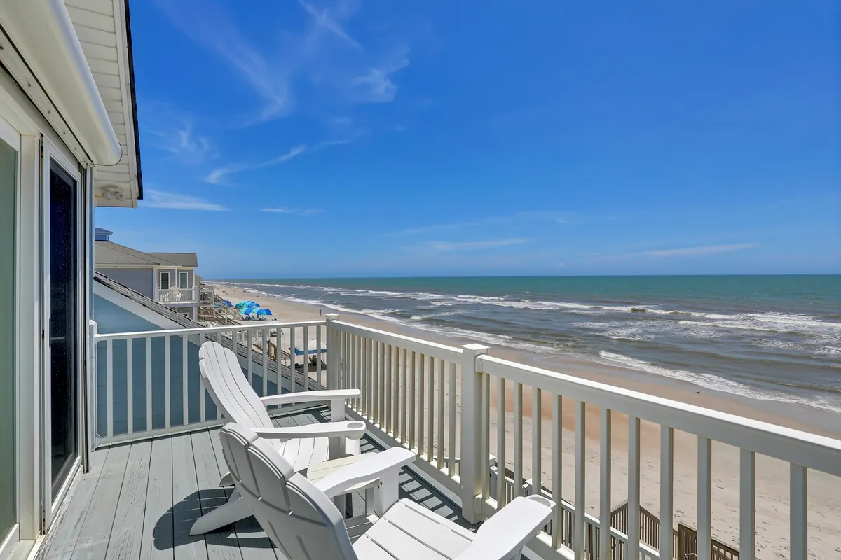 A pair of Adirondack chairs on a beach-facing deck offers a direct view of the ocean, sandy shoreline, and open sky. This inviting outdoor space is framed with natural coastal elements and suggests a quiet place for guests to relax, read, or enjoy the calming sound of the waves.