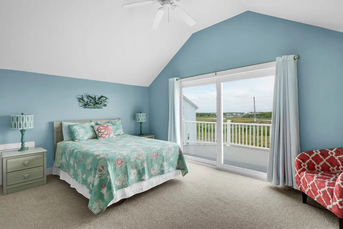 A calm and inviting bedroom with a bed featuring patterned bedding, a sliding glass door leading to a private deck, a decorative lamp, and a red accent chair. The space is styled for comfort and tranquility, inviting guests to step outside and enjoy peaceful views.